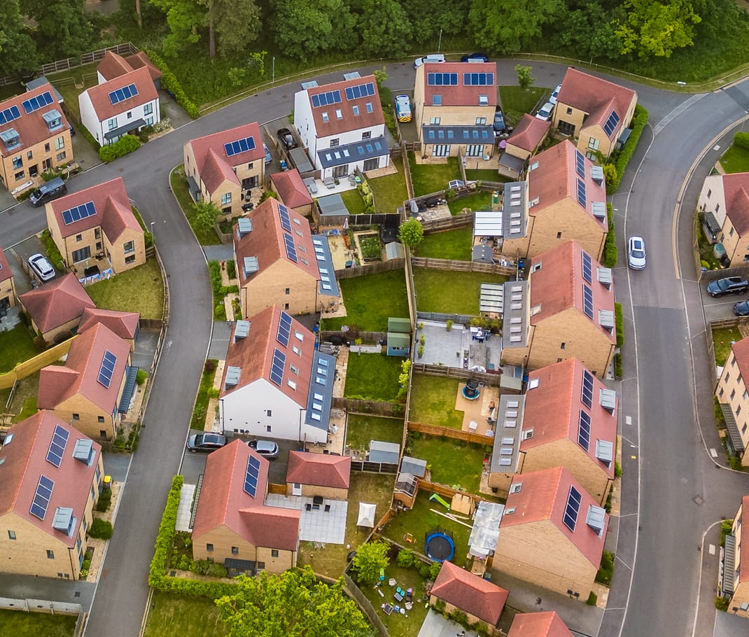 houses Aerial view of a suburban neighbourhood with rows of houses featuring red roofs and solar panels. Yards with green lawns line the winding streets.