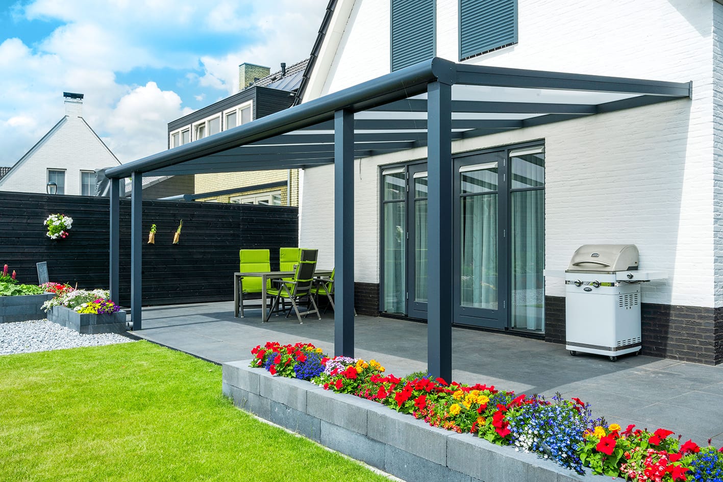 deponti-2 Modern patio with a sleek pergola, green chairs around a table, vibrant flower beds, and a barbecue grill. Bright, sunny day adding a cheerful mood.