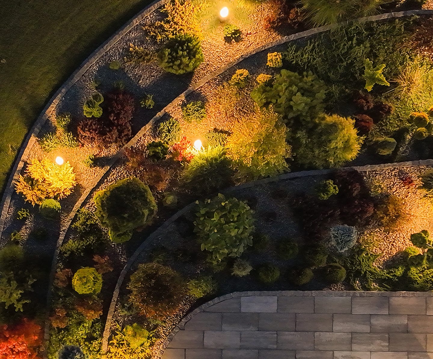 garden-aerial-view-eve Aerial view of a landscaped garden at night, featuring illuminated curved pathways and vibrant green shrubs, creating a serene and inviting atmosphere.