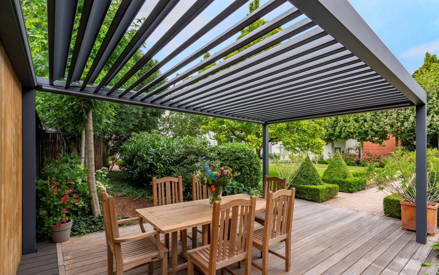 deponti A wooden patio set under a modern pergola with a slatted roof. Lush greenery surrounds the area, creating a peaceful and inviting garden atmosphere.