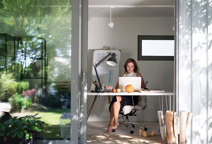 Mature woman working indoors in home office in container house in backyard. Woman working at a desk in a modern, sunlit home office. Lush garden visible outside. Calm, focused atmosphere with open laptop and office supplies.