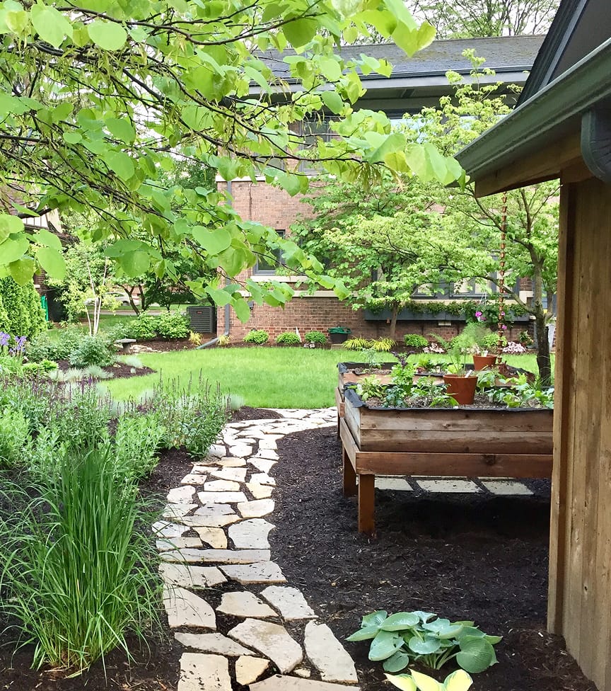 garden-path-1 Serene garden scene with lush greenery, a stone path, and a raised wooden planter. Trees frame a red-brick house in the background.