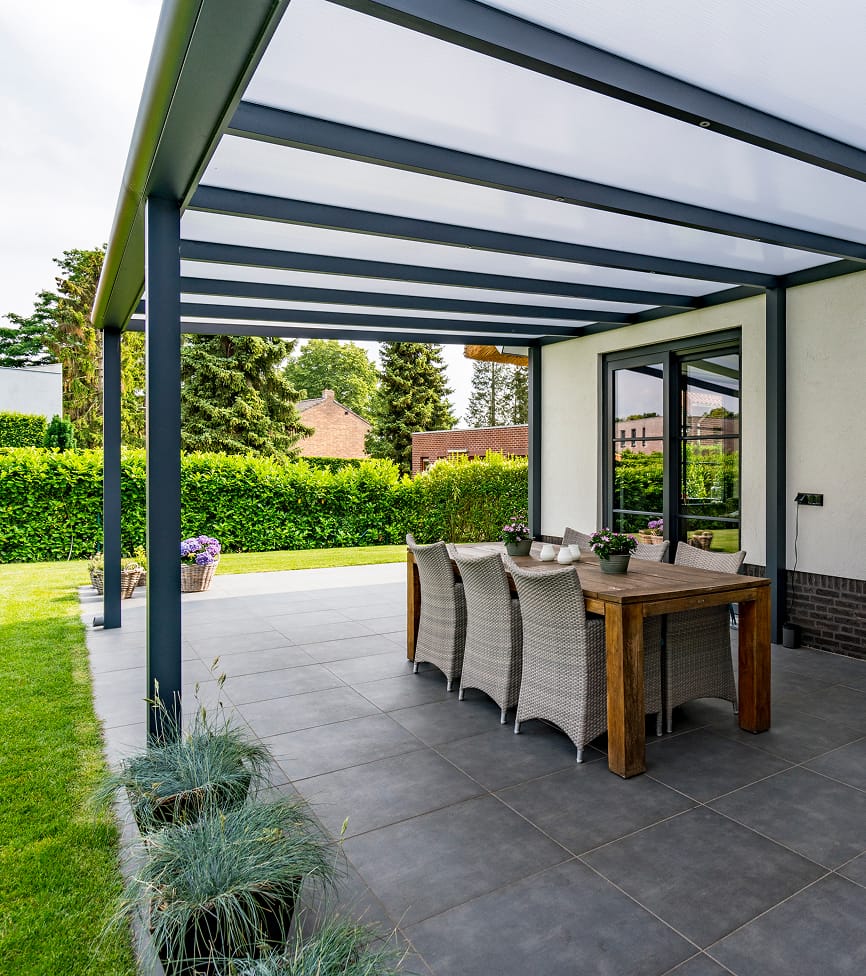 pergola-1 Outdoor patio with a modern pergola covering a wooden dining table and wicker chairs on tiled flooring. Surrounding lush greenery creates a tranquil setting.