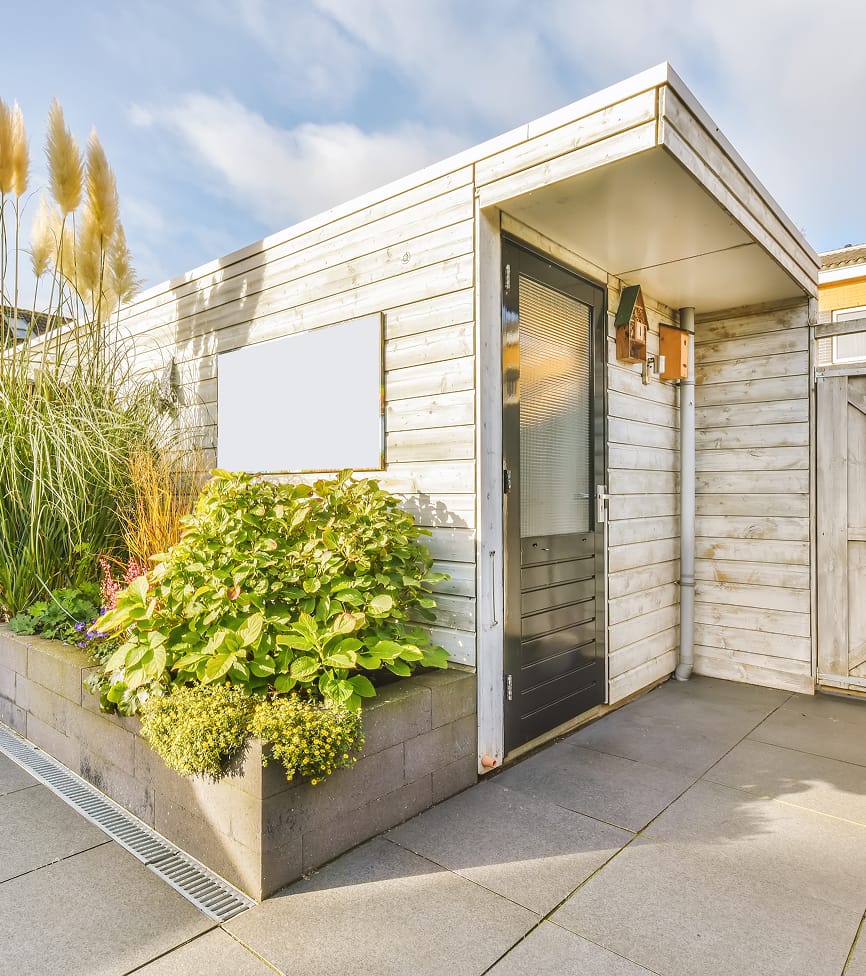 room-1 Modern wooden garden shed with black door and potted plants in concrete beds. Sunlight casts soft shadows on the stone patio, creating a serene atmosphere.